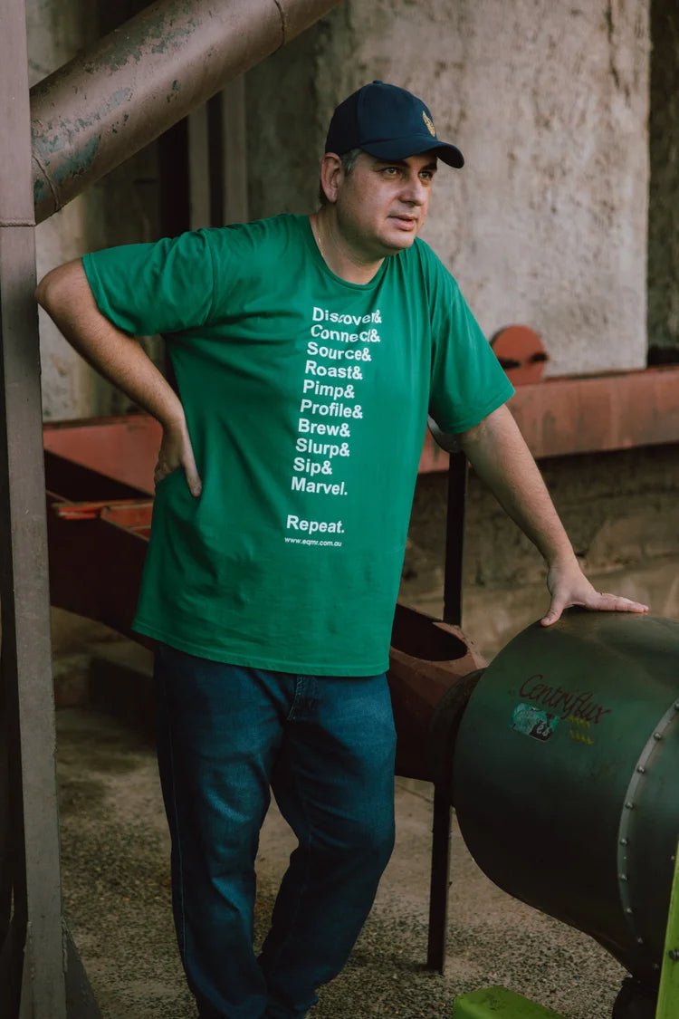 Man in green shirt, jeans, and baseball cap stands near industrial machinery.
