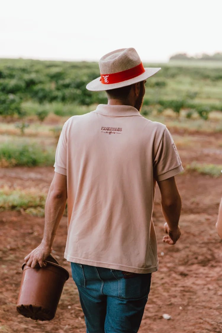 A man in a polo shirt and hat, holding a plant pot, walking in a field.