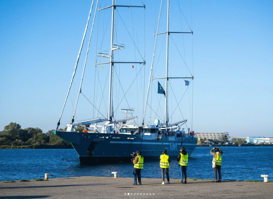 A large sailing vessel, called Anemos, with four people, three in yellow vests, standing on a concrete dock.